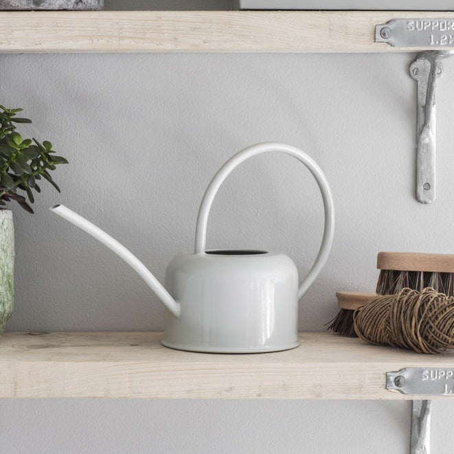 White watering can on a wooden shelf with plants and decorative items.
