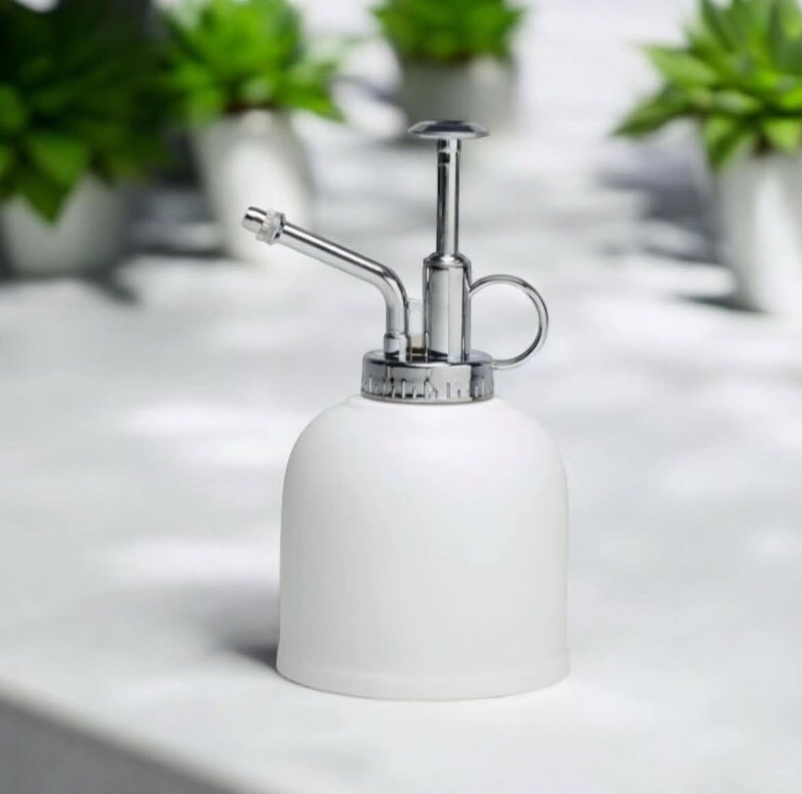 White and silver plant sprayer on a white surface with blurred green plants in the background