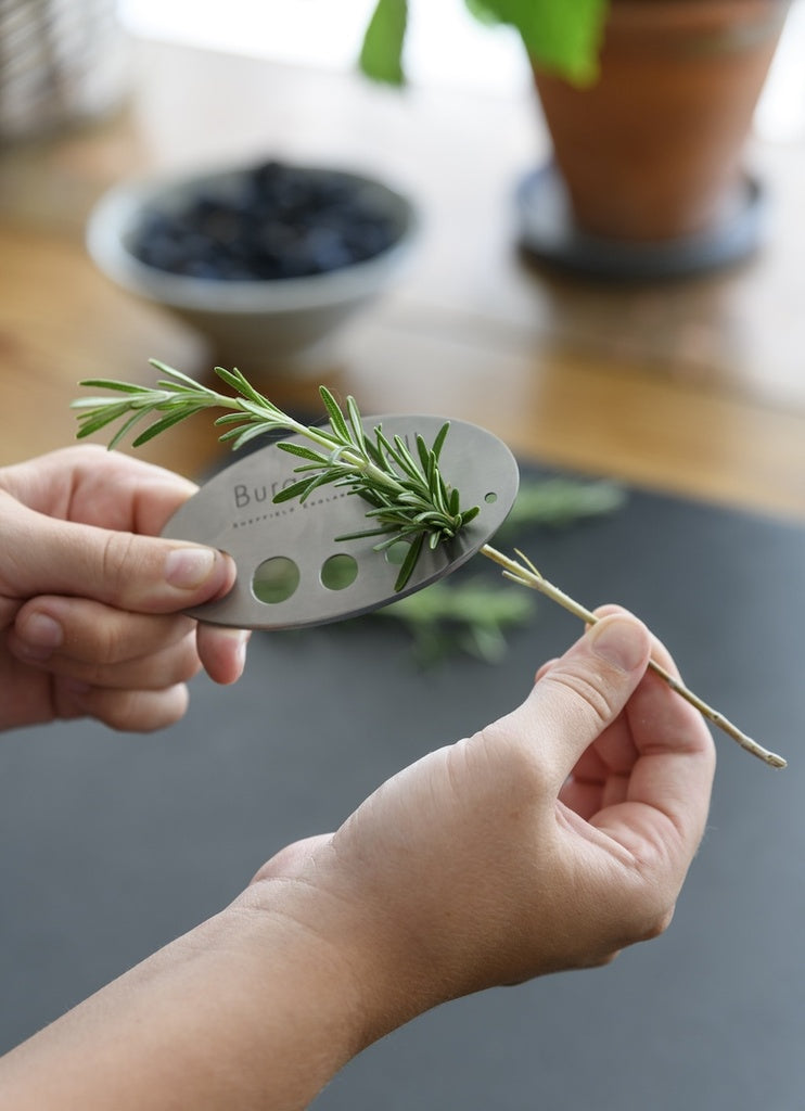 Person using a herb stripper to remove rosemary leaves from a branch on a blurred kitchen background.