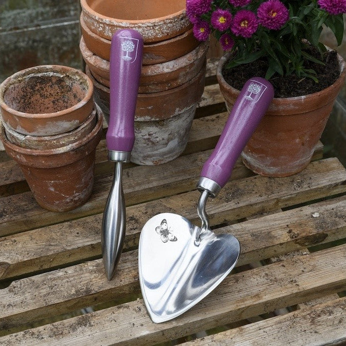 Gardening tools with purple handles on a wooden surface with potted plants.