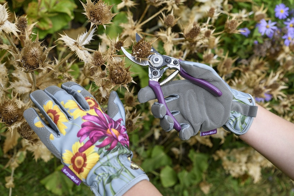 Gardening gloves with floral designs and a pair of pruning shears held by a person against a garden background.