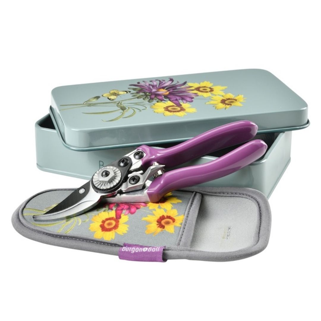 Gardening shears with purple handles in a floral-themed tin box on a white background