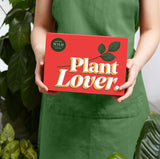 Person holding a red box labeled 'Plant Lover' with greenery in the background