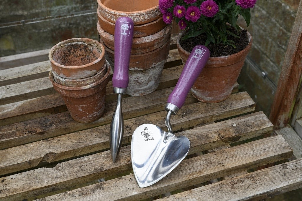Gardening tools with purple handles on a wooden surface with potted plants.