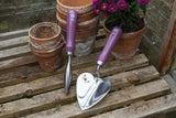Gardening tools with purple handles on a wooden surface with potted plants.