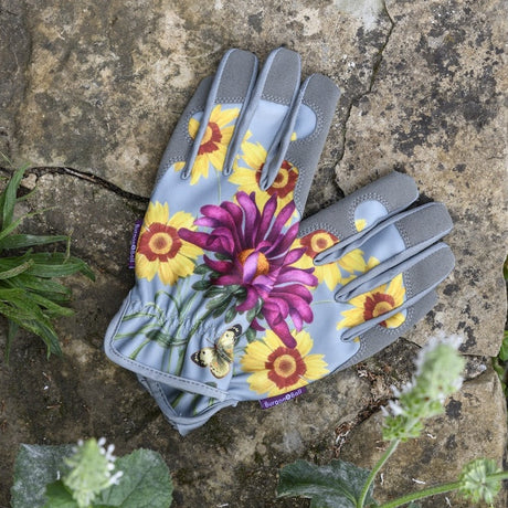 A pair of gardening gloves with a gray base and a vibrant floral print featuring yellow sunflowers and a large purple flower, laid flat on a stone surface with green plants around them.