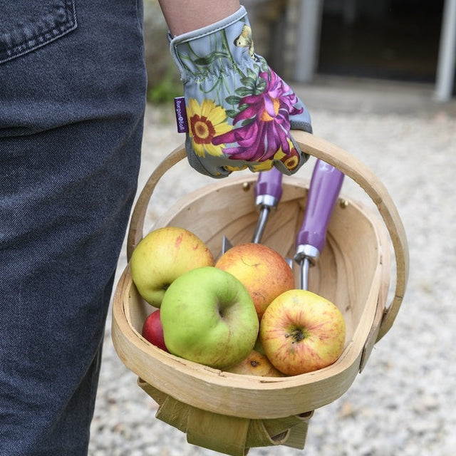 A person wearing dark jeans holds a light wood basket filled with apples and gardening tools, including a floral-patterned gardening glove.
