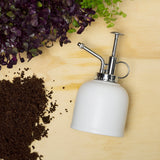 White porcelain plant mister  on a wooden surface with soil and plants.